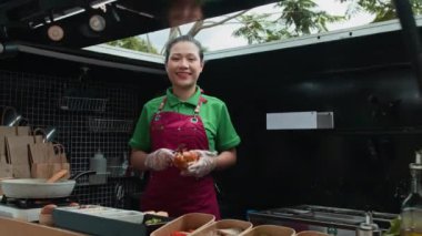 Kırmızı önlüklü genç Asyalı kadının portresi. Ekmek çöreklerinden sandviç yapıyor ve yemek kamyonunda kameraya gülümsüyor.