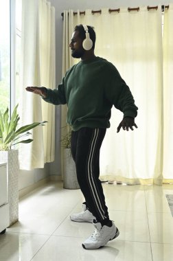 Kablosuz kulaklıklı Afrikalı Amerikalı adam evde müzik dinliyor ve dans ediyor.