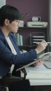 Tekerlekli sandalyedeki Asyalı kadın çalışanın modern ofiste masa başında belgelerle çalışmasının dikey görüntüsü.