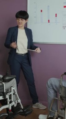 Asyalı kadın finans müdürünün, tekerlekli sandalyedeki meslektaşlarına finansal göstergeler hakkında sunum yaparken dikey çekimi
