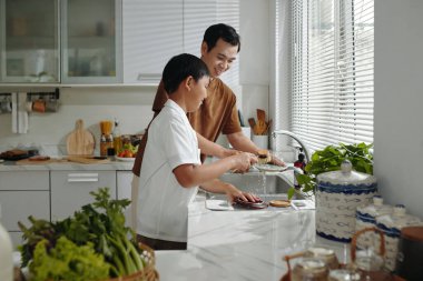 Erkek ve çocuk mutfak tezgahında yemek hazırlıyor, sebzeleri temizlemeye ve kesmeye odaklanırken etraflarında çeşitli mutfak malzemeleriyle bağlanıyorlar.