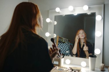 Makyaj fırçasını tutan kadın soyunma odasındaki ışıklı aynanın önünde makyaj yapıyor. Aksesuarlar ve kahve fincanı tezgahta görünür.