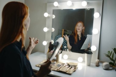 Cilt bakımını açıklayan genç bir kadın parlak ışıklı aynanın önünde elinde fırça ve kağıt tutarken. Görünür yansıma bağlılığı artırır