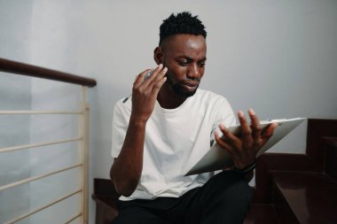 Afrika kökenli Amerikalı bir erkek, merdivenlerde otururken tabletle meşgul oluyor ve yüzüne yakın elle odaklanmış bir ifade sergiliyor.