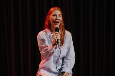 Sahnede elinde mikrofonla ayakta duran mutlu kadın stand-up komedisi yapıyor ve rutini boyunca seyircilerle takılıyor.