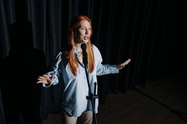 Sahnede sahneye çıkan kızıl saçlı kadın, elinde mikrofon, arka planda siyah perdeler, sahne ışıkları altında şarkı söyleyerek ya da konuşarak tutkusunu ifade ediyor.