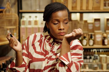 Woman with striped dress holding vintage lipstick, contemplating choices in a well-lit shop surrounded by wooden shelves and a variety of products clipart