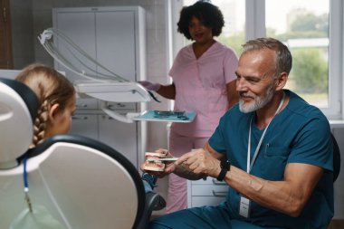 Pediatrik dişçi, diş muayenesinde asistanı olan genç bir hastayı muayene ediyor. Tıbbi tesiste hastayla uğraşırken dişçi aletini gösteriyor