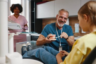 Senior dentist showing patient a denture model in modern dental office with assistant nearby. Dentist smiling and engaging with patient while demonstrating dental equipment clipart