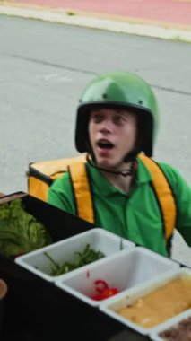 Yeşil üniformalı ve kasklı beyaz erkek kuryenin motosiklet kullanırken yemek kamyonundan sipariş verirken dikey görüntüsü.