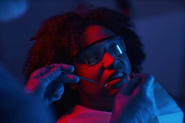 Modern klinik ortamında koruyucu gözlük takan hasta diş muayenesi yaptırıyor. Diş hekimi hastaların ağız sağlığına odaklanırken özel aletler tutuyor