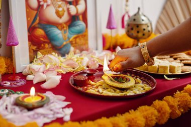 Hand performing Hindu ritual with offerings and ornamental decorations on red table. Traditional items including lamps, flowers, and sweets arranged for celebration clipart