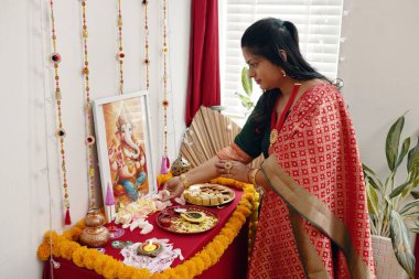 Geleneksel Hint kıyafetleri giymiş bir kadın sunağın üzerinde Tanrı Ganesh 'i sunuyor. Kültürel uygulamalara odaklanan ortamı sayısız ayin ve festival unsurları süslüyor