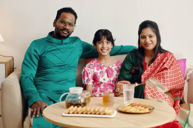 Family of three dressed in traditional attire sitting on a couch smiling together, with snacks and drinks on the table in front of them clipart