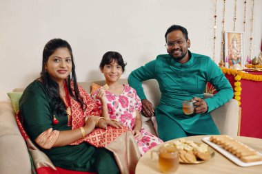 Smiling family sitting on couch enjoying snacks and drinks displaying festive decorations in background creating warm cozy atmosphere clipart