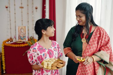 Smiling woman in traditional attire and young girl in festive dress holding trays with sweets and snacks, celebrating Indian festival with decorations in background clipart