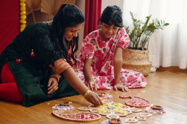 Anne ve kızı, Rangoli 'nin güzel tasarımlarını ahşap zeminde yapıyorlar. Birlikte güzel anların tadını çıkarıyorlar. Arka planda saksı bitkileri olan parlak ışıklı bir oda, aile bağlarını paylaşıyor.