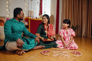 Smiling family sitting on floor and celebrating traditional festival at home with colorful decorations and intricate artwork on ground clipart