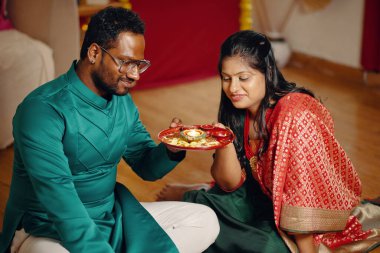 A couple partaking in cultural ceremony, holding an intricately designed plate with candle and decorations Adorned in traditional clothing, they reflect joy and commitment to heritage clipart