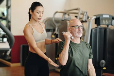 Asyalı bayan antrenör, spor salonunda halter egzersizinde yaşlı erkek müşteriye yardımcı oluyor. Forma ve formda gelişime odaklan.