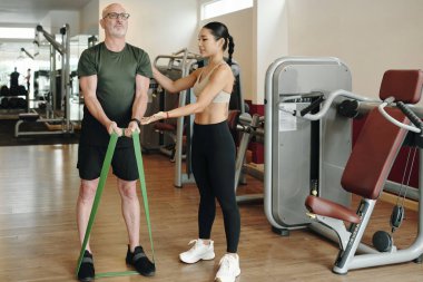 Modern spor salonu ekipmanları ve profesyonel rehberlikte dururken son sınıf öğrencilerine direnç bandı egzersizleri yaptıran kadın fitness eğitmeni de kurulumun bir parçası.