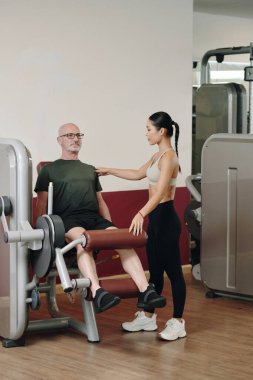 Modern jimnastik salonundaki genç bayan çalıştırıcının yardımıyla antrenman yapan yaşlı bir adam, güdümlü egzersiz seansı gösteren fitness ekipmanları kullanıyor.