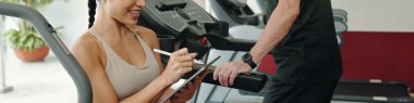 Gülümseyen antrenör elinde tablet, spor salonunda müşteri çalıştırma verilerini kaydediyor. Müşteri yanında durur, spor kıyafeti giyer, egzersiz rutininde geri bildirim alır.