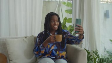 Neşeli Afro-Amerikan uzaktan kumandalı kadın yönetici kanepede kahve içerken iş arkadaşıyla telefon görüşmesi yapıyor.