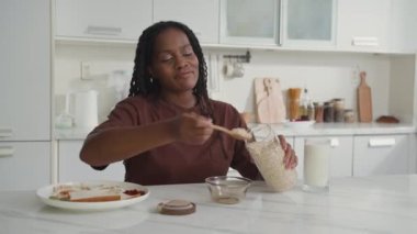 Siyahi genç bir kadın mutfakta kahvaltı hazırlarken elinde kavanozla yulaf ezmesini kaseye dolduruyor.