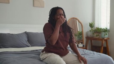 Siyah genç bir kadın uyandıktan sonra esniyor, ellerini uzatıyor ve yatak odasında başıyla dönüşümlü hareketler yapıyor.