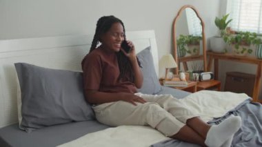 Gülümseyen siyahi genç kadın hafta sonu yatakta otururken arkadaşlarıyla telefonda konuşmaktan zevk alıyor.