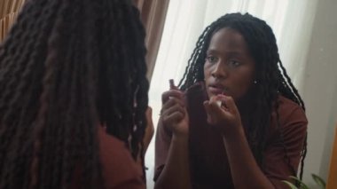 Evinde aynanın önünde dudak parlatıcısı kullanan Afrikalı Amerikalı genç bir kadının omuz manzarasından.