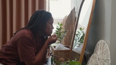 Siyahi genç bir kadının aynanın önünde makyaj masasında otururken dudaklarını pamukla temizleyişinin yan görüntüsü.