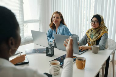 Proje fikirlerini tartışan bir grup profesyonel dizüstü bilgisayarlar, kağıtlar, fincanlar ve aletlerle çevrili modern ofiste. Takım üyeleri, birlikte çalışan tesettürlü bir kadın da dahil.