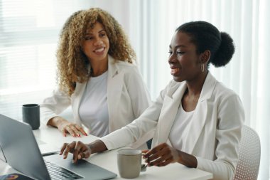 İki Afro-Amerikalı kadın, ofis masasında dizüstü bilgisayar ve kahve fincanlarıyla otururken iş konuşuyorlar. Projede işbirliği yaparken gülümseme