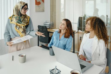 Modern ofis ortamında üç iş kadını fikirlerini paylaşıyor ve iş stratejilerini tartışıyor. Bir sohbete dahil olduklarında, odaklanırlar ve profesyoneldirler, masa etrafında işbirliği yaparlar.