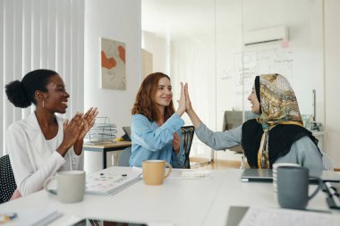 Modern ofis ortamında takım toplantısında işbirliği yapan ve başarıyı beşlik bir jest ve alkışla kutlayan üç farklı profesyonel kadın.