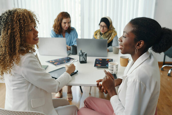 Multi-ethnic team of three women and one man collaboratively working at modern office table with various devices, engaging in conversation and sharing ideas
