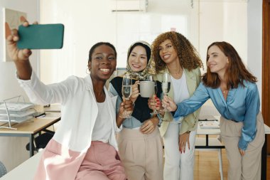 Ofiste grup selfie çekerken ve kadeh kaldırmak için kadeh kaldırırken dört farklı kadın gülümsüyor. Meslektaşlar birlikte neşeli anlar yaşıyorlar.