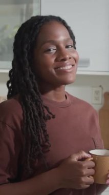 Mutfakta kahvaltı ederken kameraya bir fincan kahveyle poz veren neşeli siyah kadının dikey portresi.