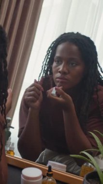 Siyahi bir kadının dudak parlatıcısı sürerken dikey çekimi. Dışarı çıkmaya hazırlanırken aynada kendine bakıyor.