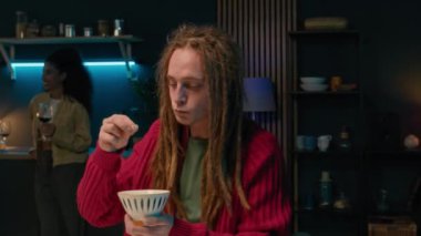 Gece partisinde elinde atıştırmalık kaseyle kameranın önünde duran uzun saçlı beyaz tenli bir gencin orta yavaş portresi. Arkadaşları bulanık arka planda sosyalleşiyor.