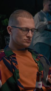 Kafkasyalı, renkli gömlek ve gözlüklü bir gencin dikey beli. Bir şişe birayla kanepede oturuyor ve arkadaşlarıyla gece partisinde akıllı telefondan kayıyor.