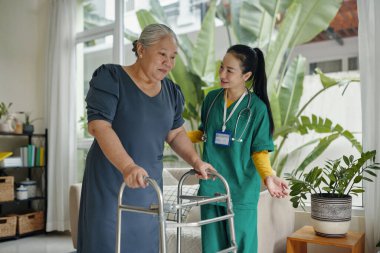 Bakıcı, yaşlı kadına odada yürümesi için yardım ediyor.