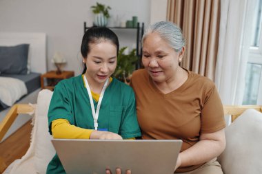 Bakıcı, yaşlı kadına odada kanepede otururken bilgisayar kullanmayı öğretiyor.