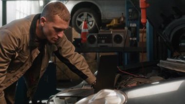 Garajda bozulma nedeni için araba parçalarını incelerken beyaz erkek tamircinin dizüstü bilgisayar kullanırken orta boy fotokopi çekimleri.