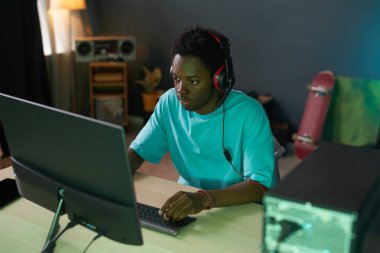 Young gamer in headphones playing vr game on computer while sitting in his room clipart