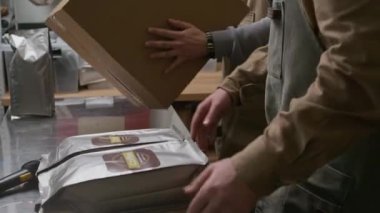 İki beyaz erkek işçiyi, fabrikada satışa hazırlanırken taze kavrulmuş kahveyle poşetleri kutuya koyarken çek.