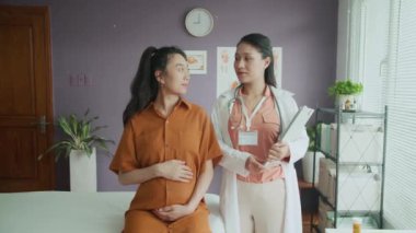 Beyaz üniformalı neşeli Asyalı kadın doğum uzmanı ve klinikteki check-up sırasında kameraya poz veren hamile anne portresi
