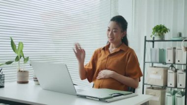 Orta ölçekli, Asyalı hamile iş kadınının ofis masasındaki videoyu izlerken dizüstü bilgisayarın önünde oturması.